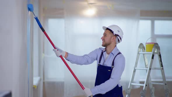 Male Painter in Uniform and Helmet Makes Repairs Indoors and Paints Walls Using Construction Roller alt