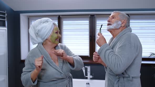 Elderly Grandmother with Facial Green Mask and Grandfather with Shaving Foam Dancing in Bathroom alt