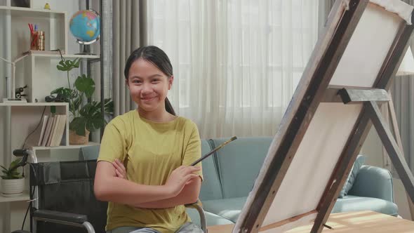 Kid Girl In Wheelchair Holding Paintbrush And Cross His Arms And Smile While Painting On The Canvas alt