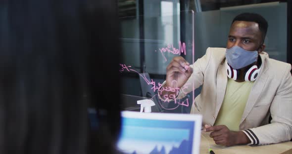 African american man wearing face mask writing on glass board at modern office alt