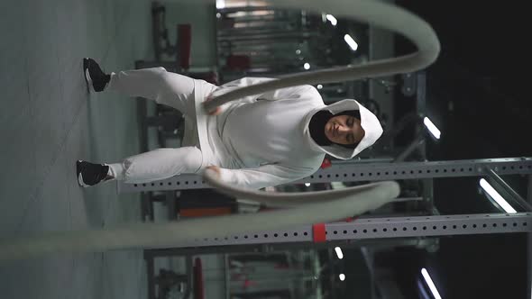 Muslim Athlete Trains in the Gym Performing Crossfit Exercises with Combat Ropes and Wears a Sports alt