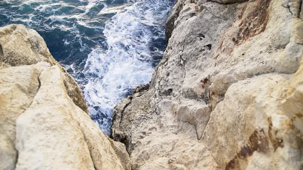 Looking down at blue ocean sea water waves crashing on steep rocky tan cliffs, Malta, zoom out alt