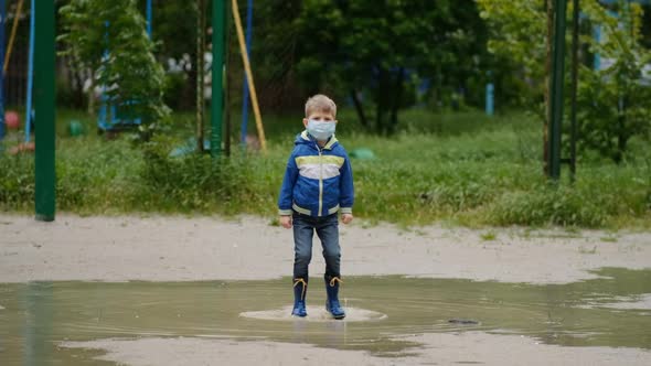A small boy in a medical mask and rubber boots jumps through puddles on a spring day after a rain.