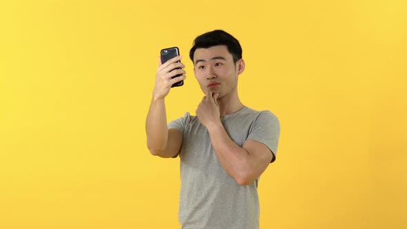 Portrait of smiling young Asian man taking selfie on mobile phone against yellow background