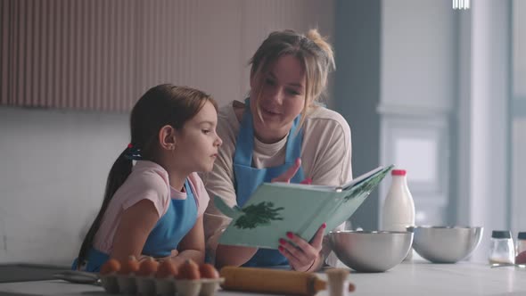 Woman and Her Little Daughter are Reading Cookbook Kitchen of House in Sunday Mother is Teaching Her alt