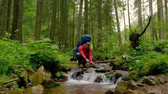 A Bearded Man with a Backpack Drinks Water From a Mountain Stream alt