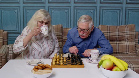 Smiling Senior Couple Grandfather Grandmother Resting on Sofa Drinking Coffee Playing Chess at Home alt