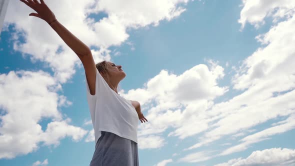 Young Happy Woman Raises Hands in Background of White Clouds and Bright Blue Sky alt