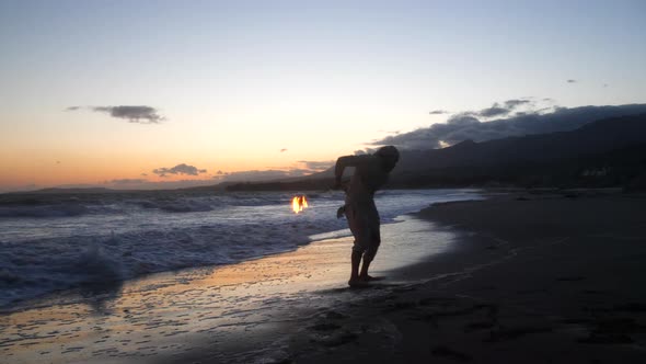 A man in silhouette dancing and spinning a burning fire staff on the beach at sunset with flames and alt