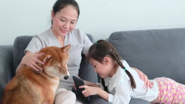 Asian mother and daughter sit on sofa with playing tablet and enjoy with dog in living room alt