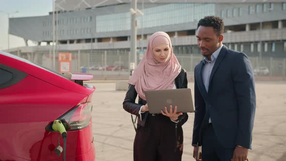 Female Dealer in Hijab Using Laptop While Showing Luxury Electric Car to Male alt