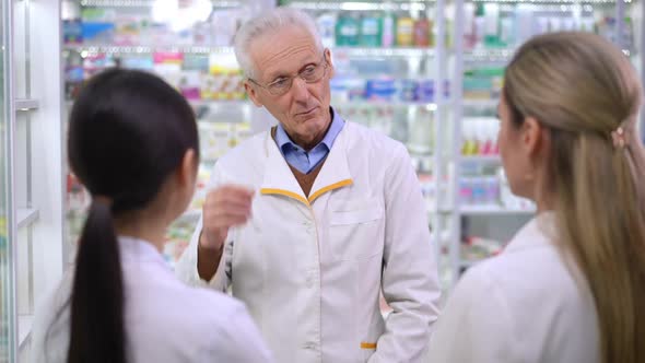Senior Man in Eyeglasses Talking with New Employees Standing at Shelves in Pharmacy alt