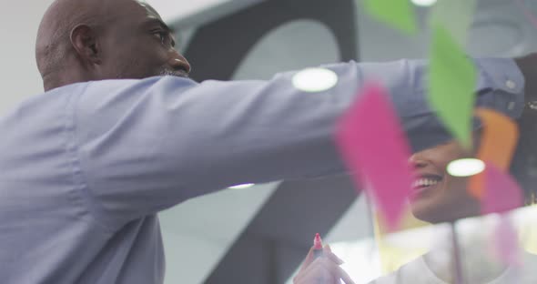 Diverse businessman and businesswoman brainstorming and smiling by transparent board with memo notes alt