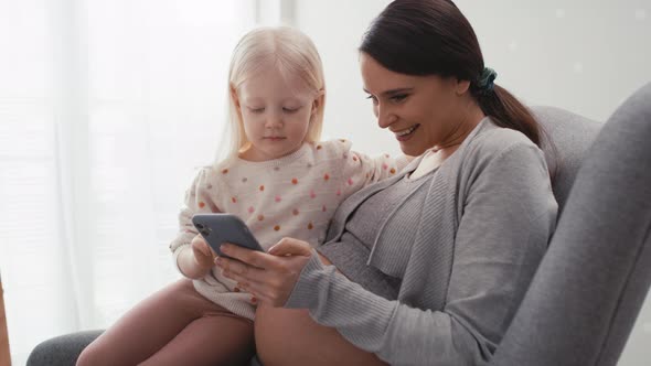 Caucasian woman in advanced pregnancy and her elementary aged daughter using mobile phone at home. S alt