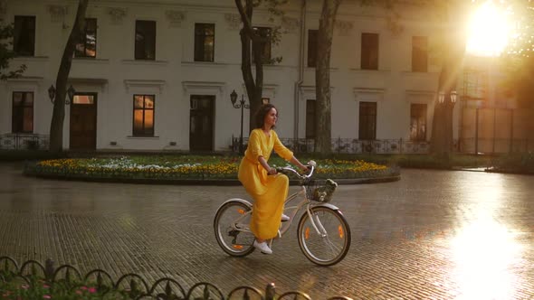 Young Woman Riding a City Bicycle with a Basket and Flowers in the City Center During the Dawn alt