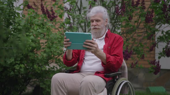 Positive Handicapped Greyhaired Caucasian Man Talking Using Video Chat on Tablet Sitting in alt