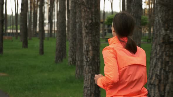 Brunette female in orange jacket running on footpath in the city park alt
