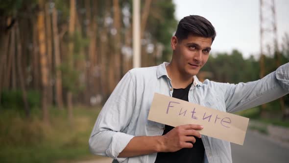 Handsome Young Positive Man Hitchhiking with Future Banner on Suburban Road at Forest alt
