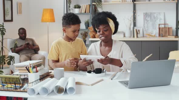 Afro-American Woman Working at Home and Helping Kid with Game on Tablet alt