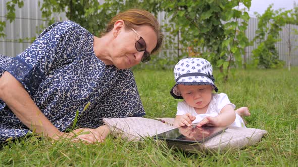Kid with Nanny Watching Cartoons. Cartoons on Tablet Watched Outdoor. Girl Lying on Grass. Childhood alt