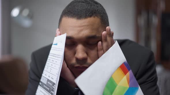 Closeup of Overwhelmed Exhausted African American Man Sighing Holding Graphs and Diagrams Sitting in alt