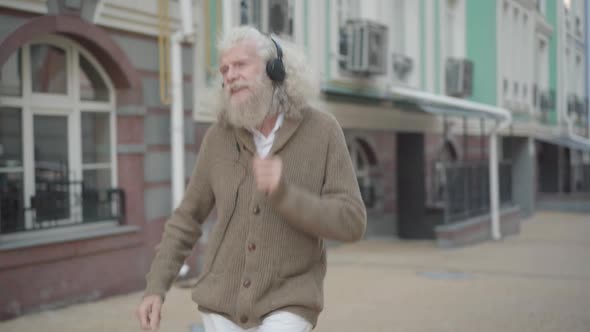 Carefree Caucasian Old Man in Headphones Dancing on City Street. Portrait of Relaxed Happy Senior alt