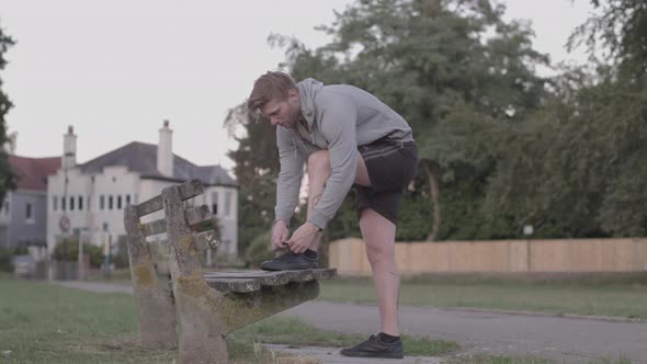Young Athletic Man Ties His Shoes on Bench in Park Before He Sets Off on Run - Ungraded alt