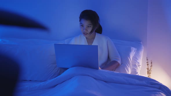 young asian female wearing casual dress sit on bed with meeting online via laptop computer