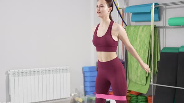 a Young Woman in a Tracksuit Does Squats with Gymnastic Elastic Bands alt
