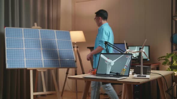 Asian Man Walks Into The Office That Has Solar Cell Next To The Laptop Showing Wind Turbine alt