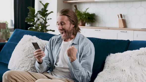 Happy Joyful Young Caucasian Guy in a Stylish Denim Shirt Sits on the Sofa in the Living Room Holds alt