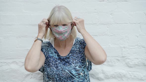 Elderly blond woman puts on floral face mask with a happy smile, safety concept for old people alt