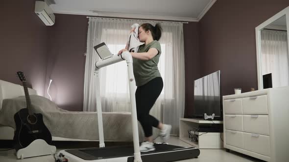 Chubby female is training at home, doing cardio fitness exercises on treadmill alt