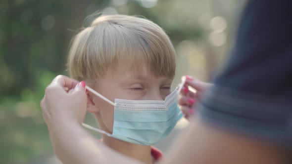Close-up of Cute Boy with Female Caucasian Hands Putting Face Mask on His Face. Charming Caucasian alt