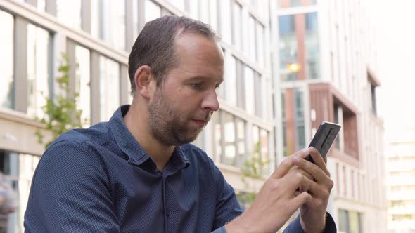 A Caucasian Man Holds a Smartphone Looks Around  Side Closeup  Office Buildings in the Background