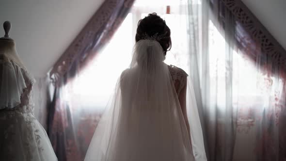 Unrecognizable Bride in White Veil Walks to Window Past Wedding Dress alt