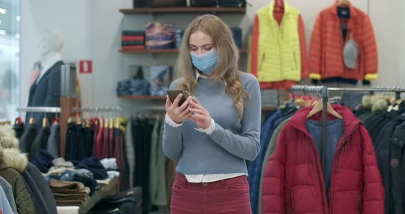 Young Caucasian Woman in Covid-19 Face Mask Using Smartphone in Clothing Shop. Portrait of Positive alt