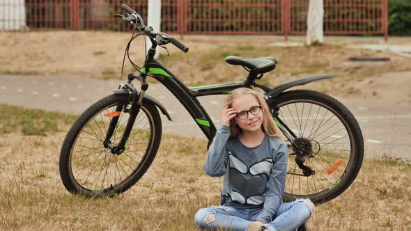 Blonde Little Girl Posing with Her Bicycle in Summer Sitting on the Grass alt