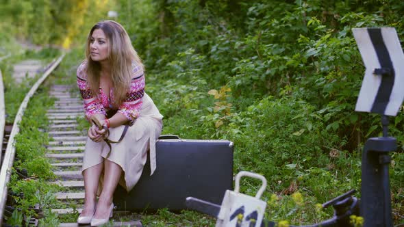 Waiting Woman Sitting on Suitcase on Railway in Summer Forest. Tourist Woman on Traveling Suitcase alt