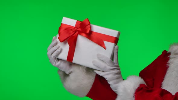 Side View of Santa Claus in White Gloves and a Red Suit Twirls a Gift Box in His Hands alt