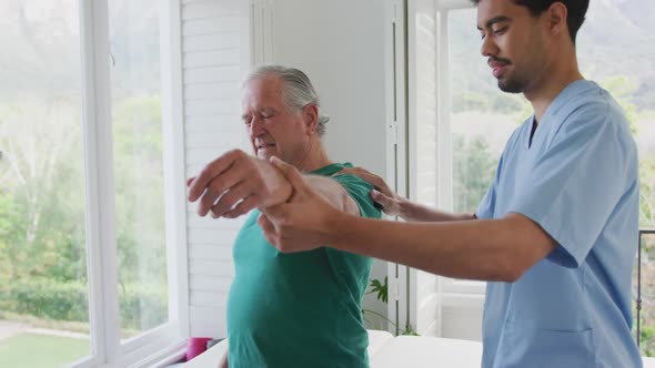 Biracial male physiotherapist slowing moving hand of senior man grimacing with pain at nursing home alt