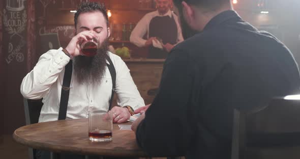 Two Bearded Men in a Bar Drinking Socializing and Start Playing a Card Game alt