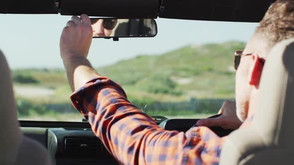 Caucasian man in sunglasses sitting in car adjusting rearview mirror on sunny day at the beach alt
