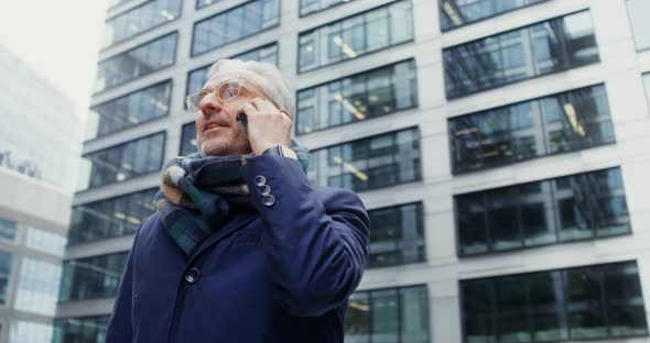 A Grayhaired Man Uses a Mobile Phone Standing in Business Center of the City alt