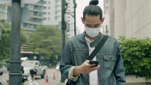 Asian man wearing a surgical mask and using a smartphone while walking on the street.