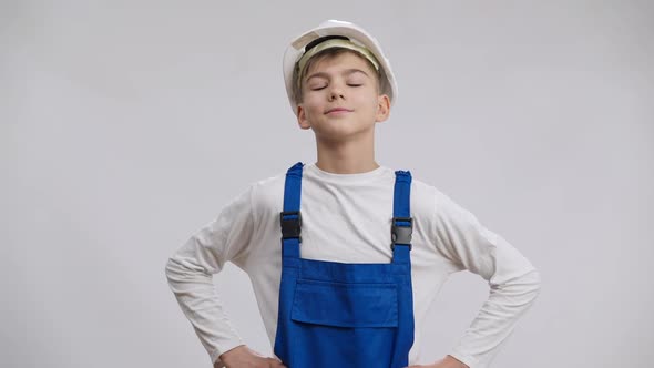 Caucasian Boy in Hard Hat and Overalls Putting Hands on Hips Posing at White Background alt