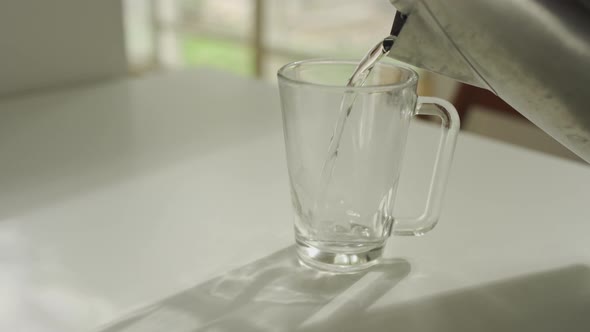 Hot water being poured into a glass mug from metal kettle in apartment alt