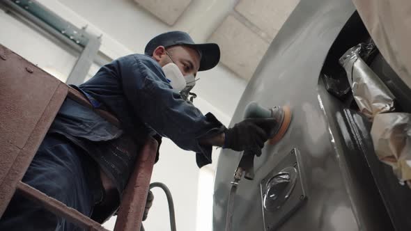 A Closeup View of a Worker in Overalls and a Respirator is Grinding the Paintwork of the Car Using a