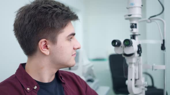Young Confident Patient Sitting at Refractometer Looking Back at Equipment Turning Smiling alt
