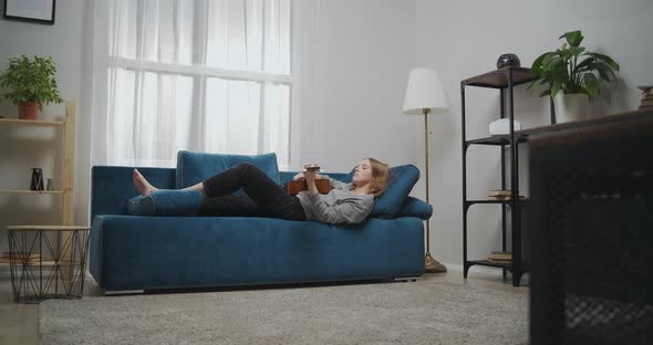 Young Girl in Casual Clothes Lies Barefoot on a Sofa in a Modern Living Room alt